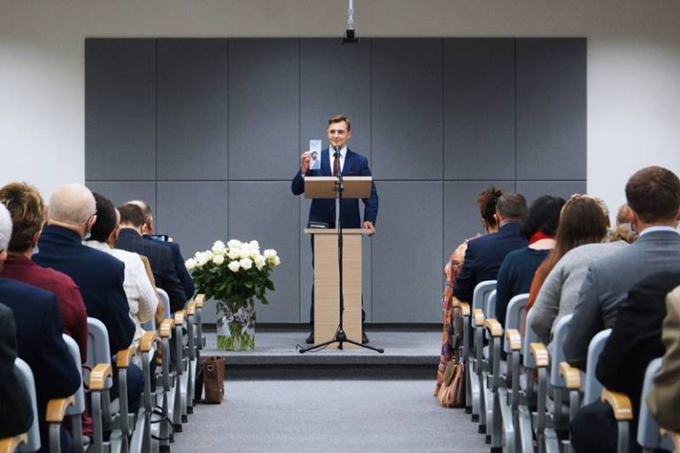 W oddali mężczyzna za mównicą przemawia do słuchaczy siedzących w ławkach