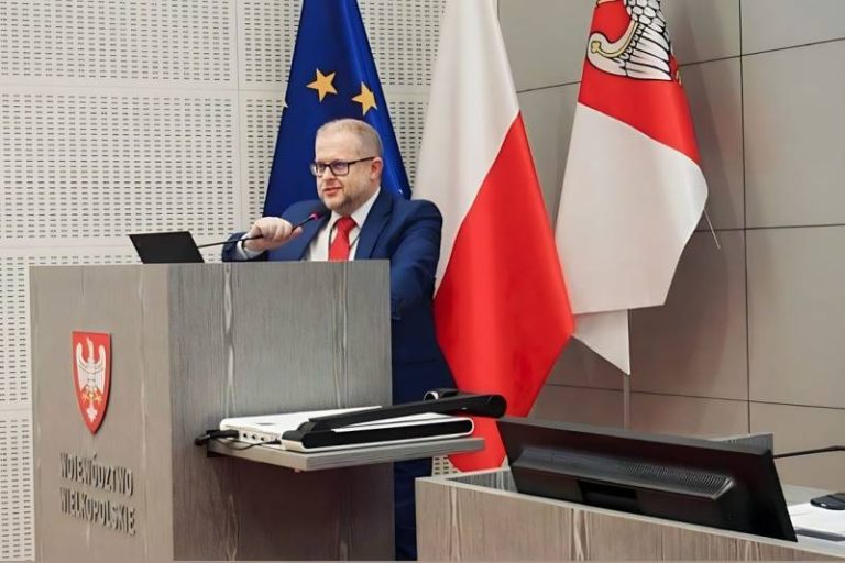 Dariusz Młynarczyk przemawia z mównicy na sali sesyjnej Urzędu Marszałkowskiego w Poznaniu; w tle flaga europejska, polska oraz Wielkopolski