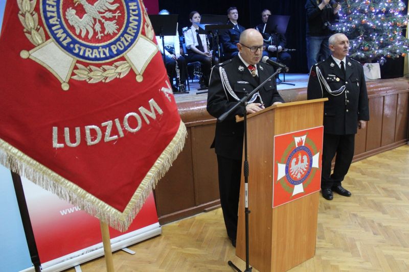 Przemawia z mównicy prezes OSP w powiecie tureckim Grzegorz Ciesielski. Na pierwszym planie sztandar strażacki